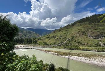 A river flows at the feet of a mountain, white clouds against blue skies hang above, a bridge is stretched across the river.
