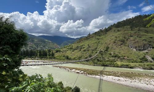 A river flows at the feet of a mountain, white clouds against blue skies hang above, a bridge is stretched across the river.