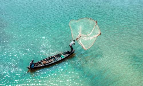 Urban communities including fishing communities face climate risks. A fisherman on a boat casts a net upon wide waters.