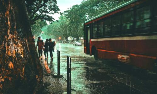 A group of people walking down a sidewalk next to a bus as it rains in Mumbai. Photo by Sadiq Sonalkar/Unsplash.